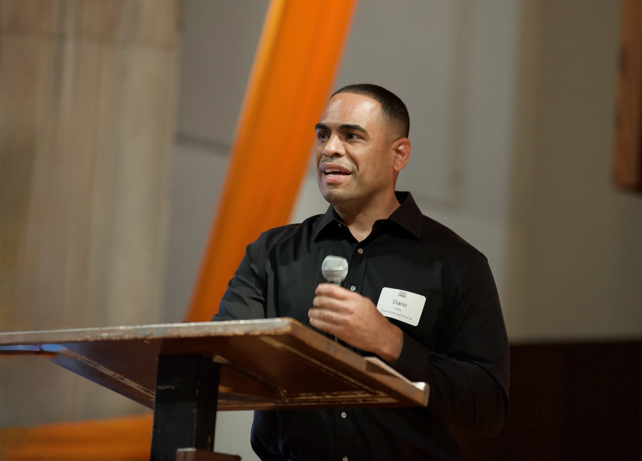 Man with a short haircut wearing a black shirt, standing at a wooden podium, holding a microphone, and speaking at an event.