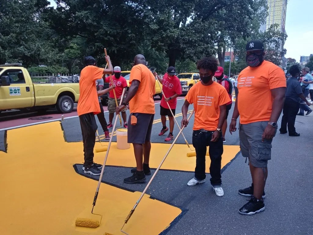 Group of people painting a yellow crosswalk on a city street, wearing matching orange shirts and masks.