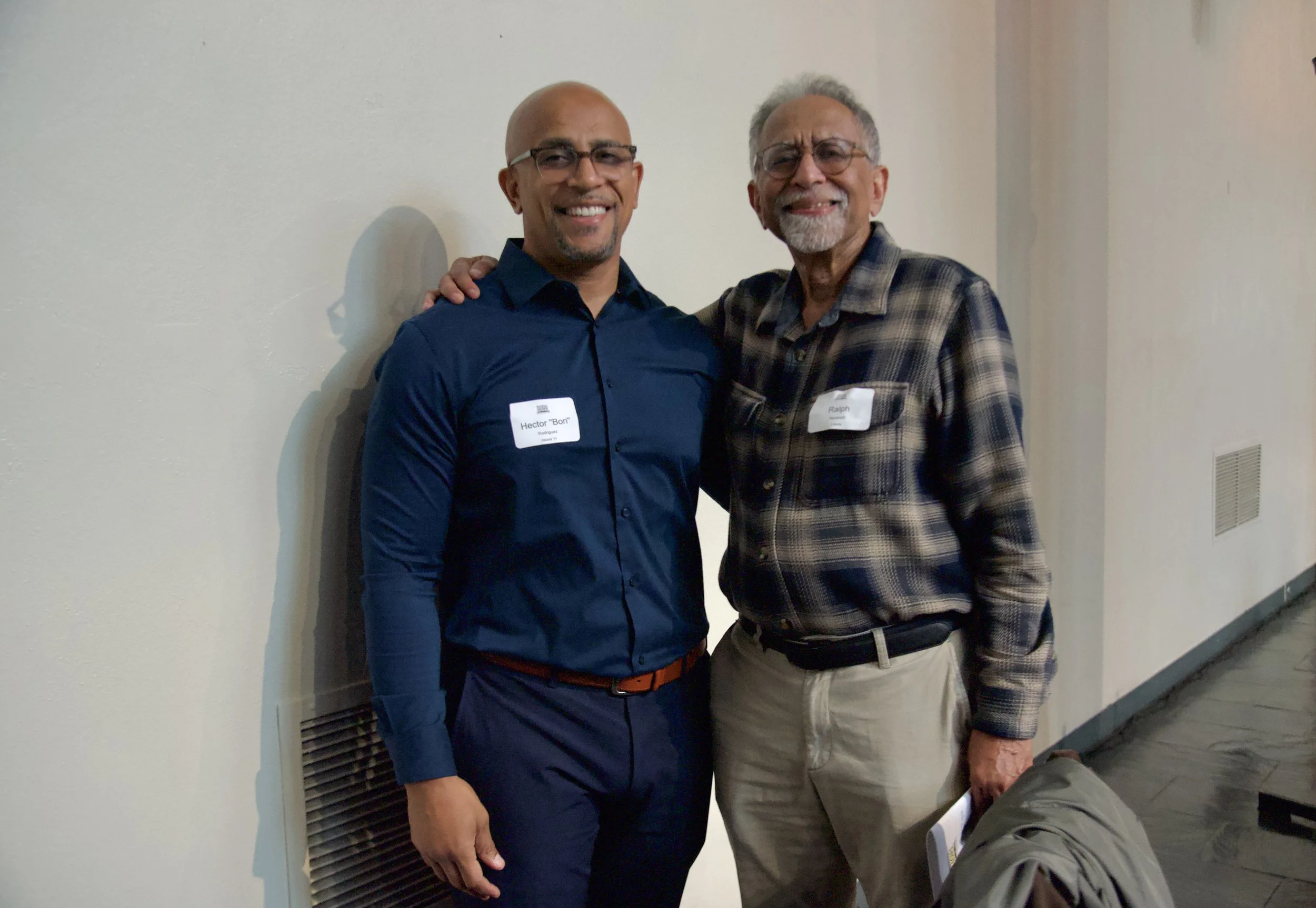 Two men smiling and standing close together indoors, one with a name tag reading 'Hector 'Bon' Rodriguez' and the other with a name tag reading 'Ralph', wearing glasses.