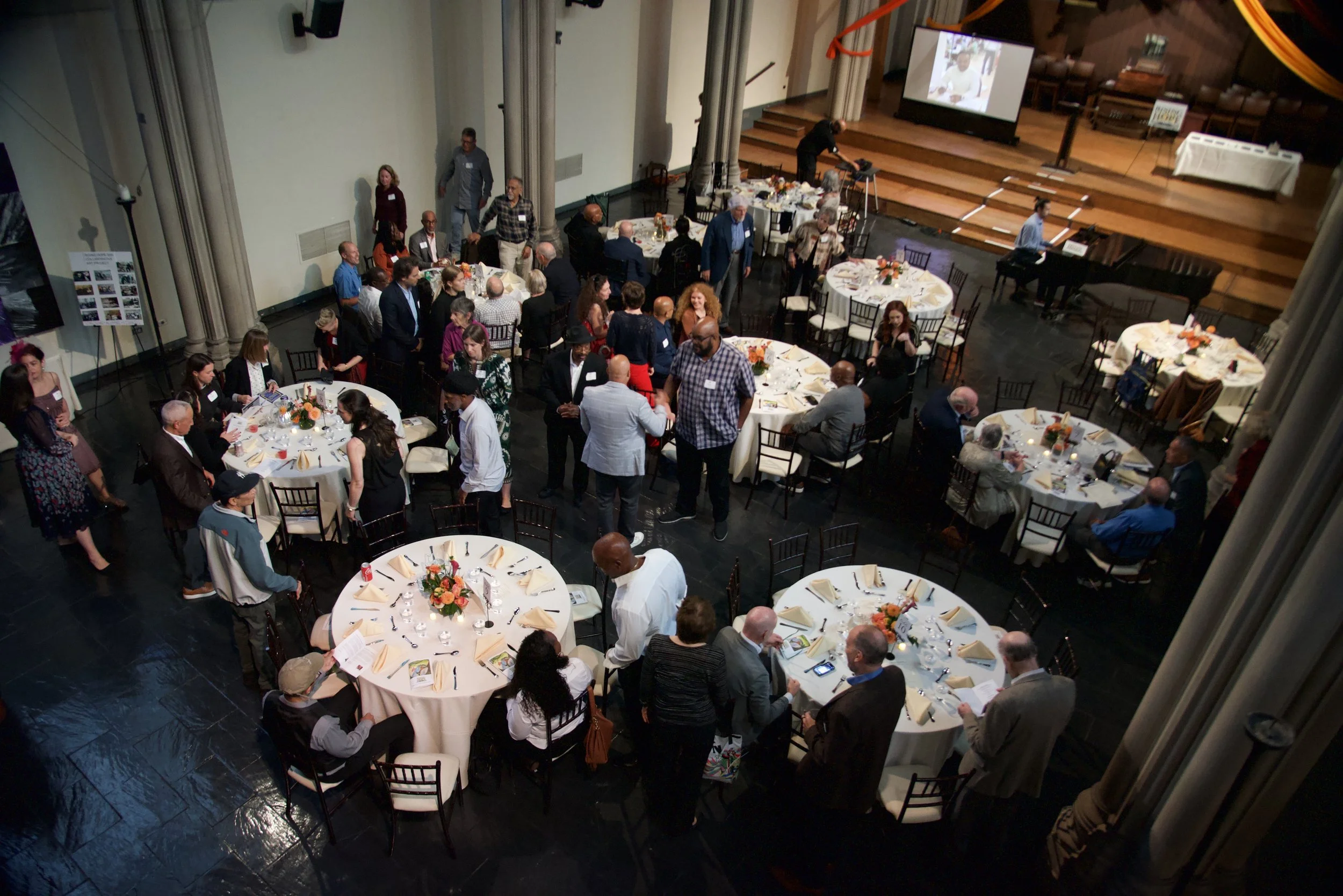 A large group of people gathered around round tables with white tablecloths, orange flower centerpieces, and place settings, in a hall with a stage and a screen displaying a photo of a person. Some individuals are seated, while others are standing an