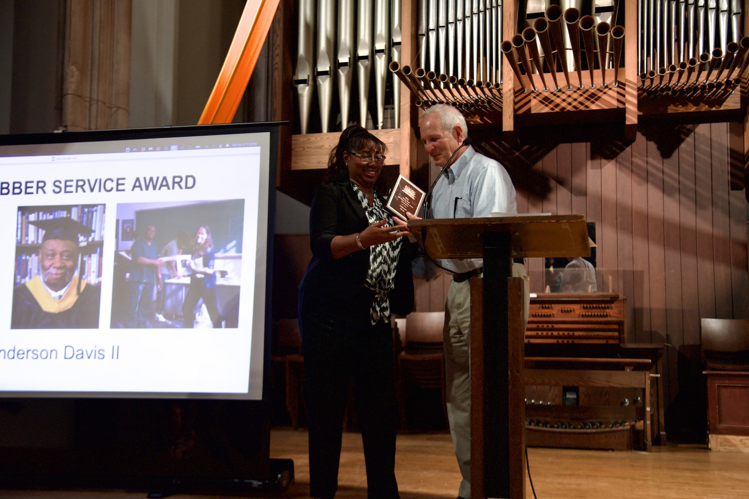 A woman receiving an award from a man at a ceremony. The background shows a large pipe organ and a wooden wall. A presentation slide with images and text is partially visible on the left.