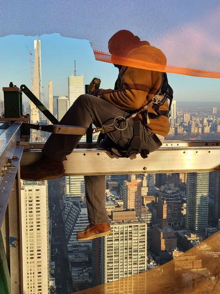 A construction worker sits on a steel beam high above a cityscape, wearing a safety harness and helmet, surrounded by skyscrapers in a high-rise building.