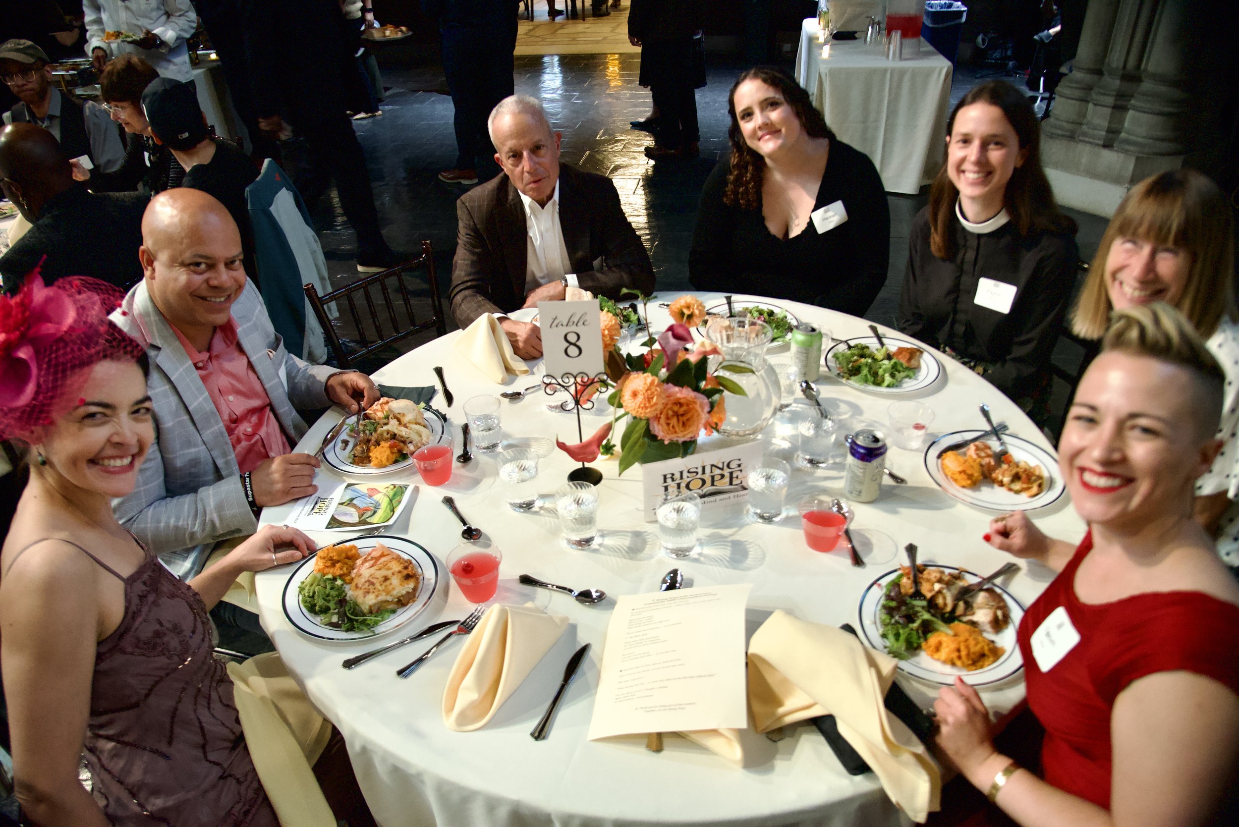 People sitting around a wedding reception table with food and drinks, smiling and enjoying the event.