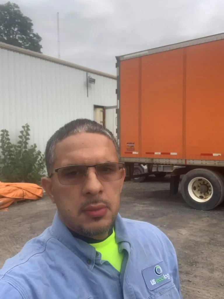 A man wearing glasses and a blue collared shirt taking a selfie outdoors near a large orange truck and a white building with some trees in the background.