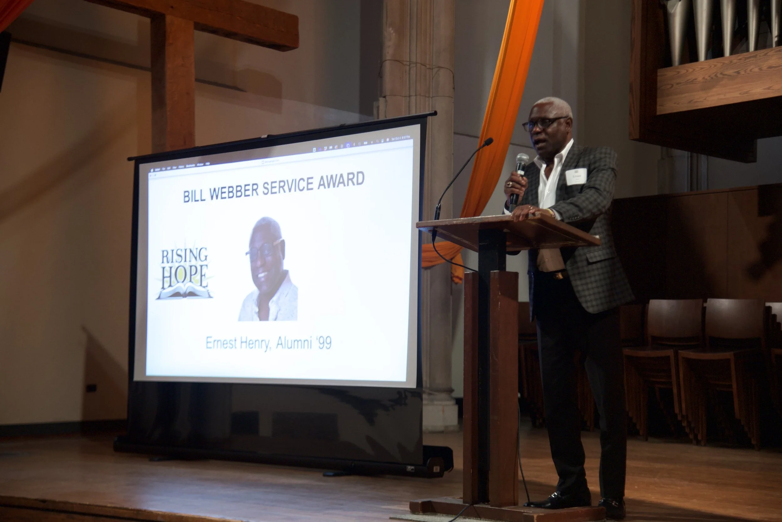 A man standing at a podium giving a speech, with a large screen behind him displaying a presentation titled 'Bill Webber Service Award' and a photograph of an individual named Ernest Henry, Alumni '99.