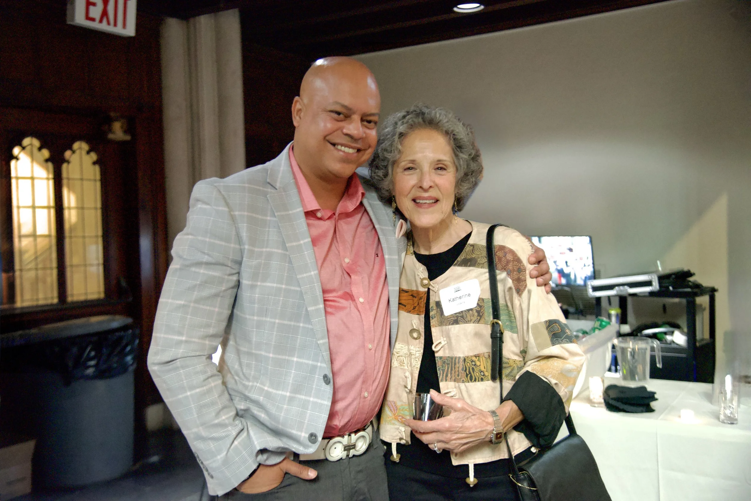 A man in a light gray checkered blazer and pink shirt standing next to an older woman with curly gray hair wearing a colorful jacket and a name tag. They are smiling and posing together indoors, possibly at a social event or gathering.