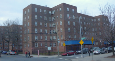 Multi-story brick apartment building with a blue awning, parking lot, and cars outside, in an urban setting.
