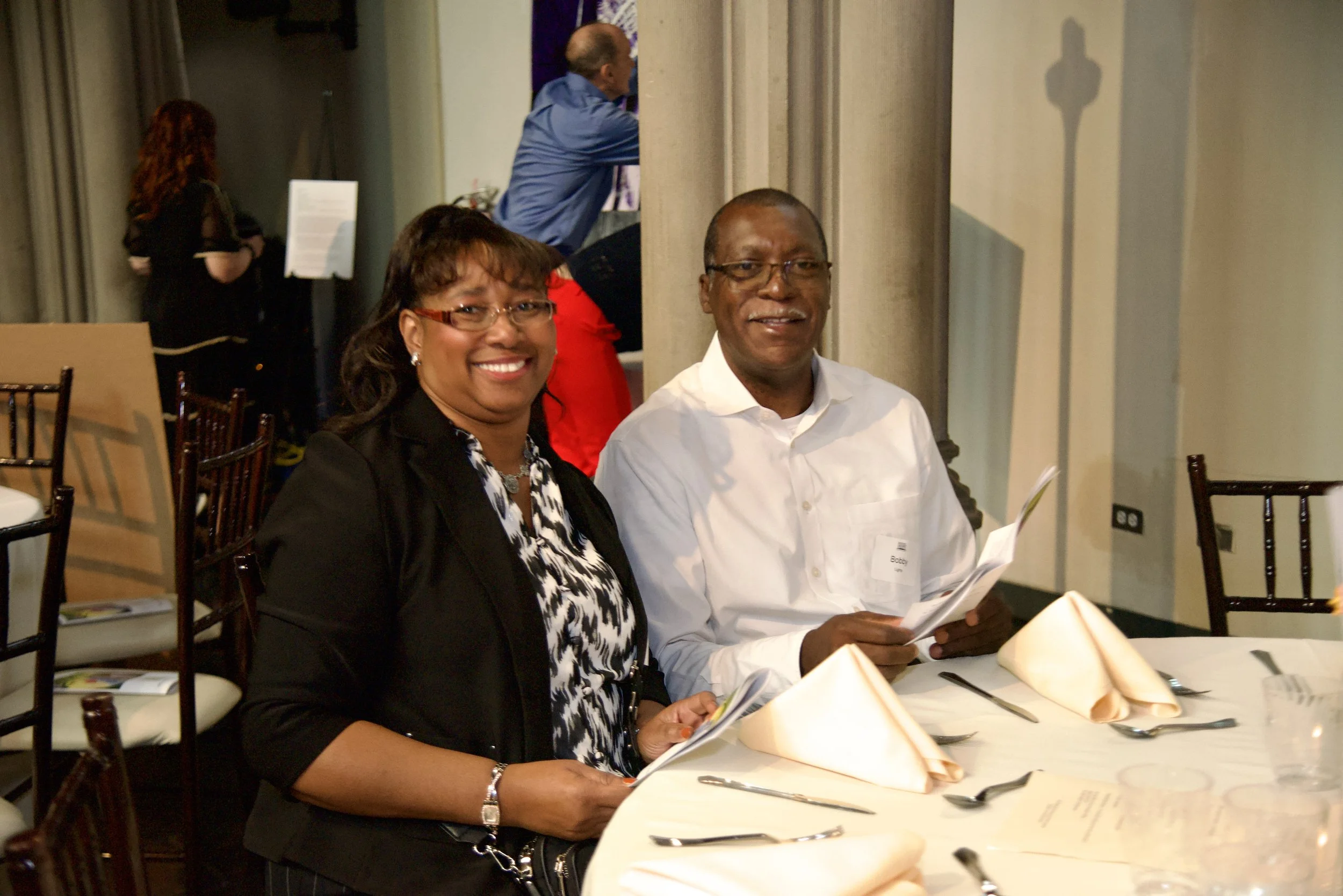 A man and a woman sitting at a restaurant table, smiling, with folded napkins, utensils, and menus in front of them. In the background, a woman with red hair and a black dress and a man in a blue shirt are talking.