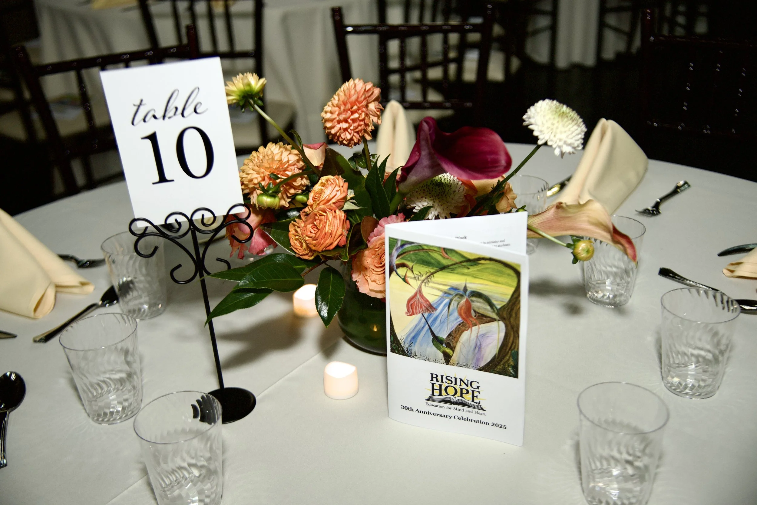 A round table decorated with a floral centerpiece, a table sign indicating table 10, and a program booklet titled "Rising Hope" for a 30th anniversary celebration. The table is set with clear glass cups, silverware, and folded beige napkins.