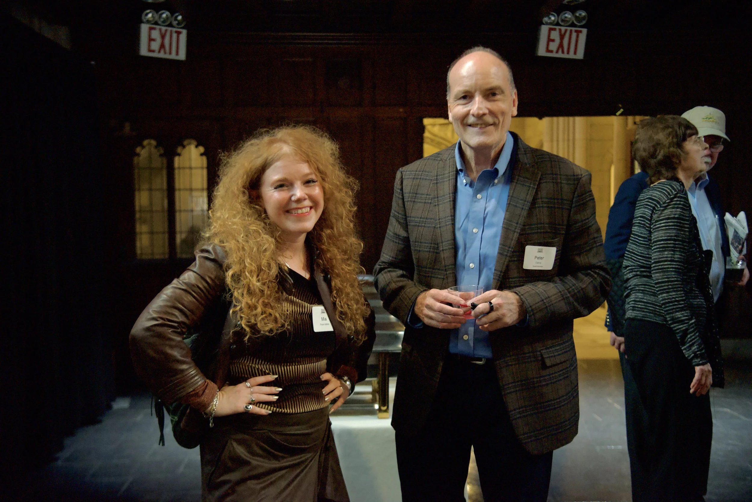 A woman with long, curly red hair and a man with a bald head and gray beard smiling at an event. The woman is wearing a leather jacket and the man is wearing a plaid blazer. They are indoors with other people in the background.