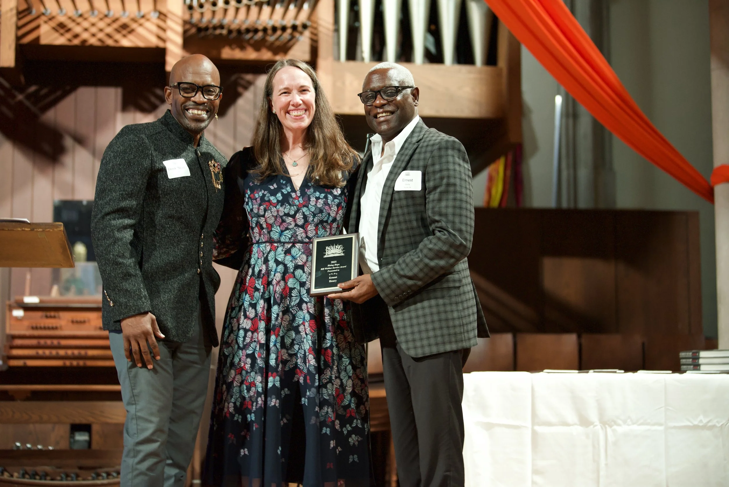 Three people standing close together, smiling, with a woman in a floral dress holding a plaque, in an indoor setting with wooden decor and a staircase in the background.