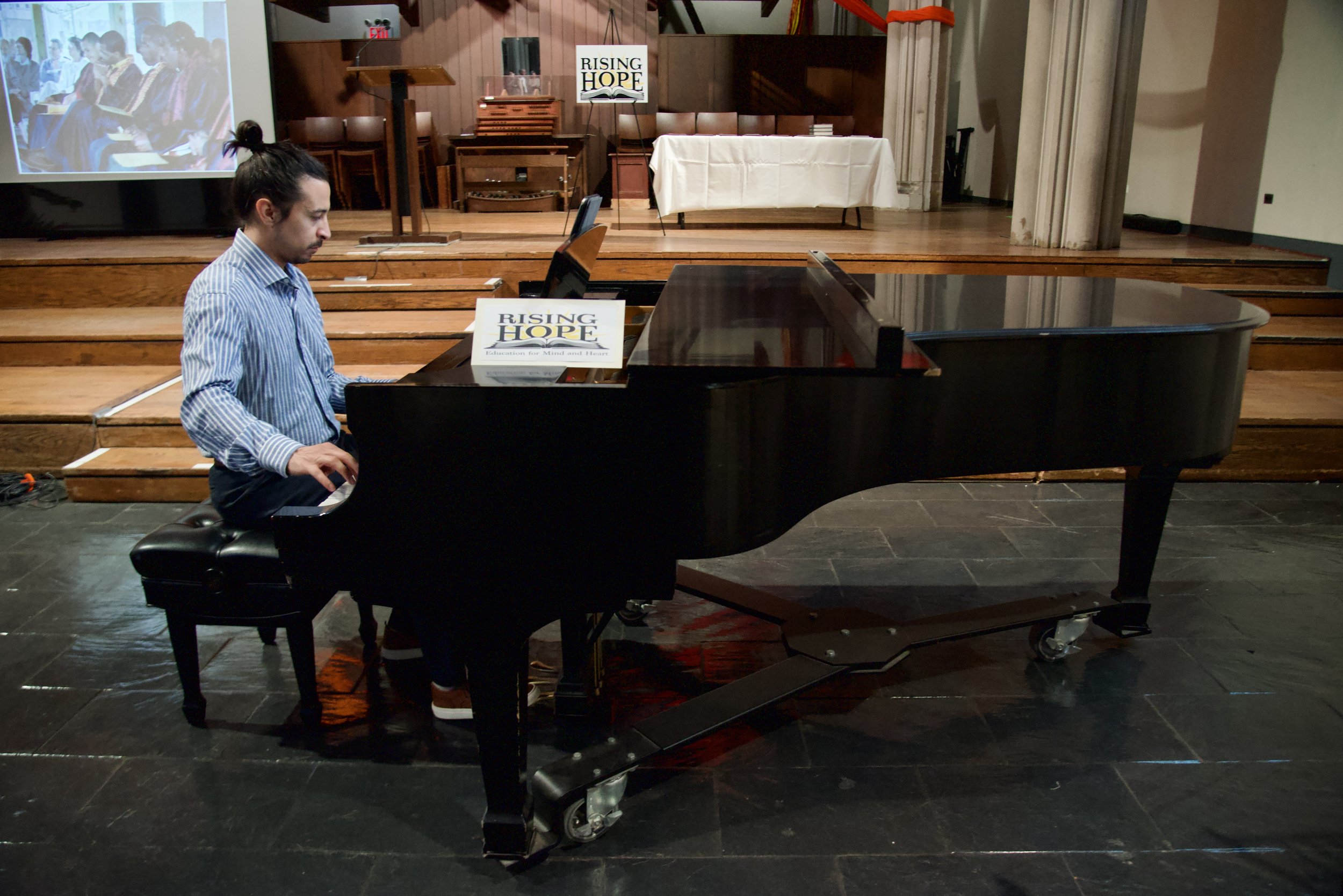 A man with long hair tied in a bun playing a black grand piano inside a church or auditorium with wooden floors and a stage. The stage has a sign that reads 'Rising Hope,' a table covered with a white cloth, a podium, and a large screen displaying an