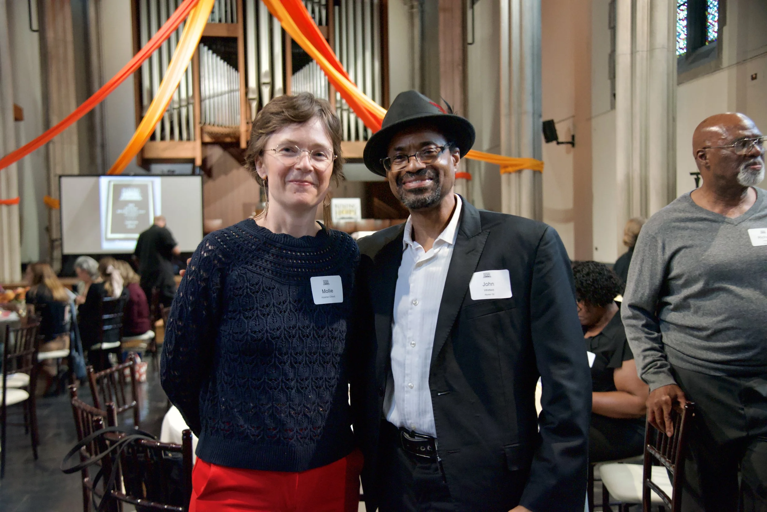 A woman and a man posing together at an indoor event, with the woman wearing a navy blue knitted top and red pants, and the man wearing a black suit, white shirt, and a black hat. They are smiling and looking at the camera, with a crowd and a pipe or