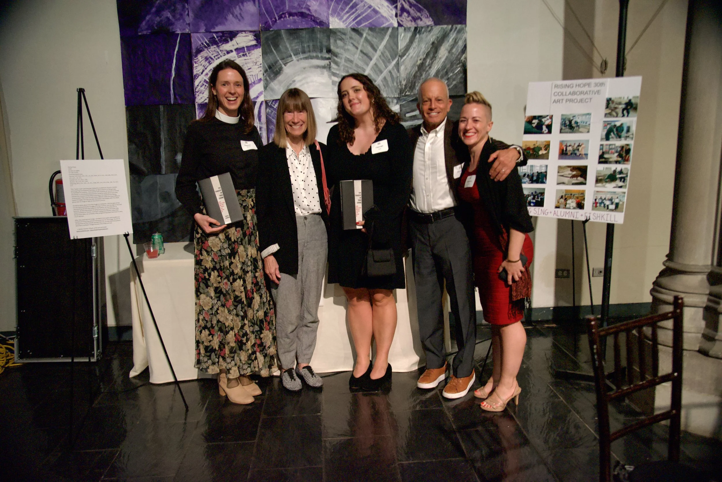 Group of five people standing together at an art event, smiling for the camera with abstract purple and gray artwork behind them and informational posters on easels to their sides.