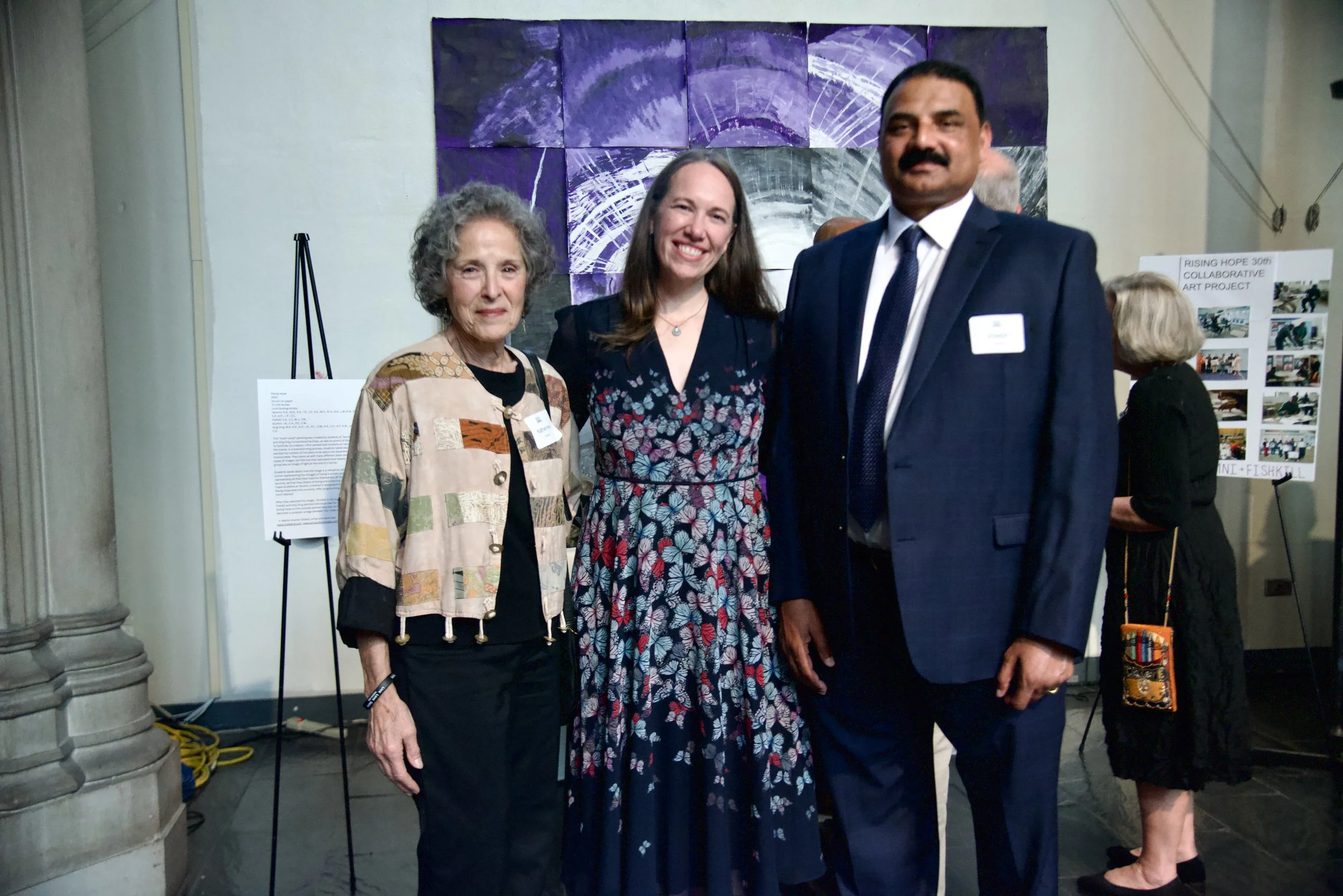 Three women and one man standing together at an art event, with purple abstract artwork on the wall behind them.