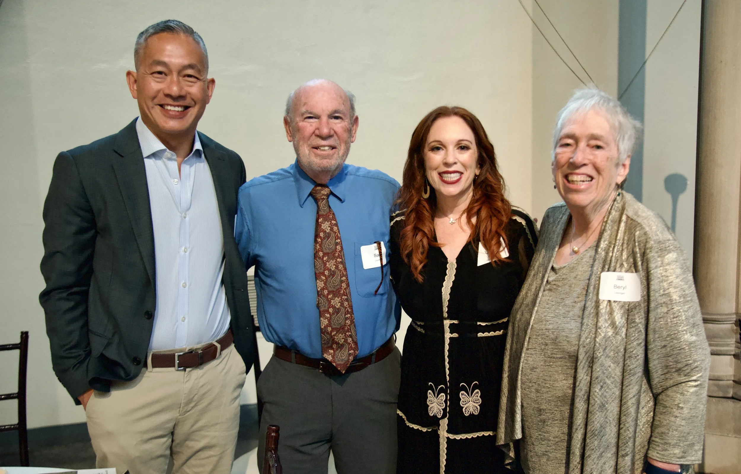 Four people standing together at an indoor event, smiling at the camera. The group includes two men on the left, a woman with red hair in the center, and an older woman with short white hair on the right. They are dressed in business casual attire.