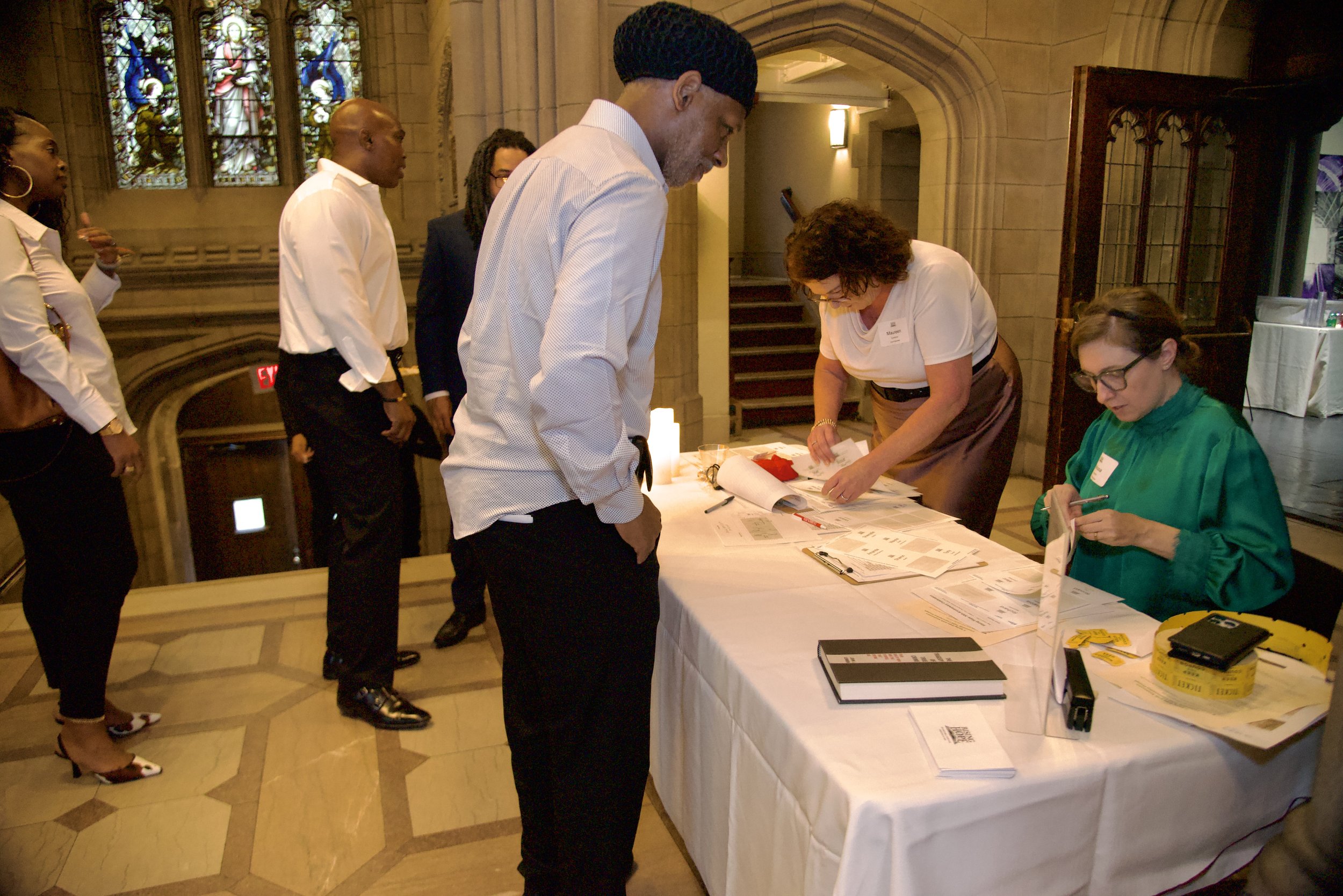 People standing in line at a registration table inside a church with stained glass windows, signing documents and handling paperwork.
