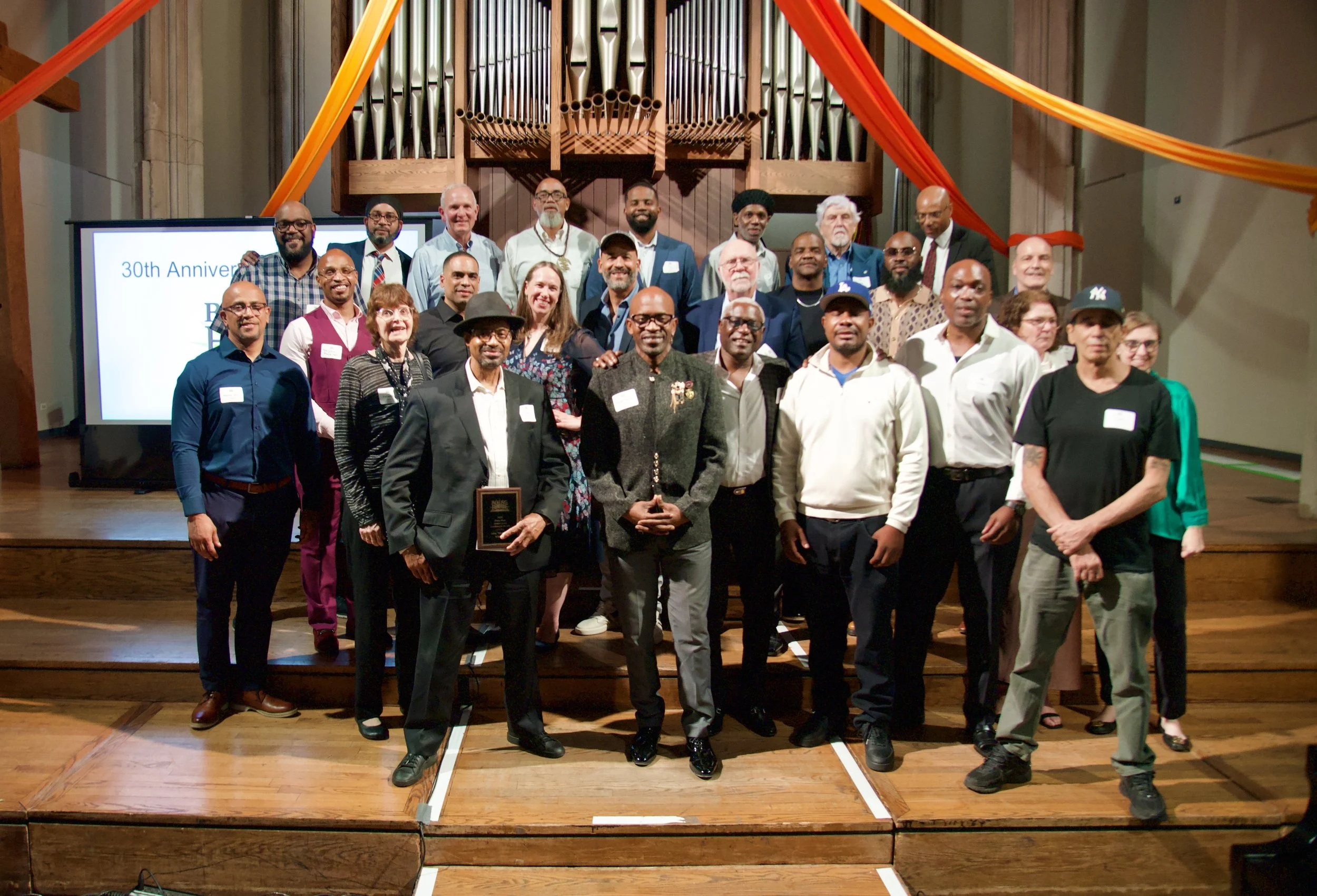 Group of people standing on a stage in front of a pipe organ, celebrating an anniversary, with colorful drapery decorations overhead.