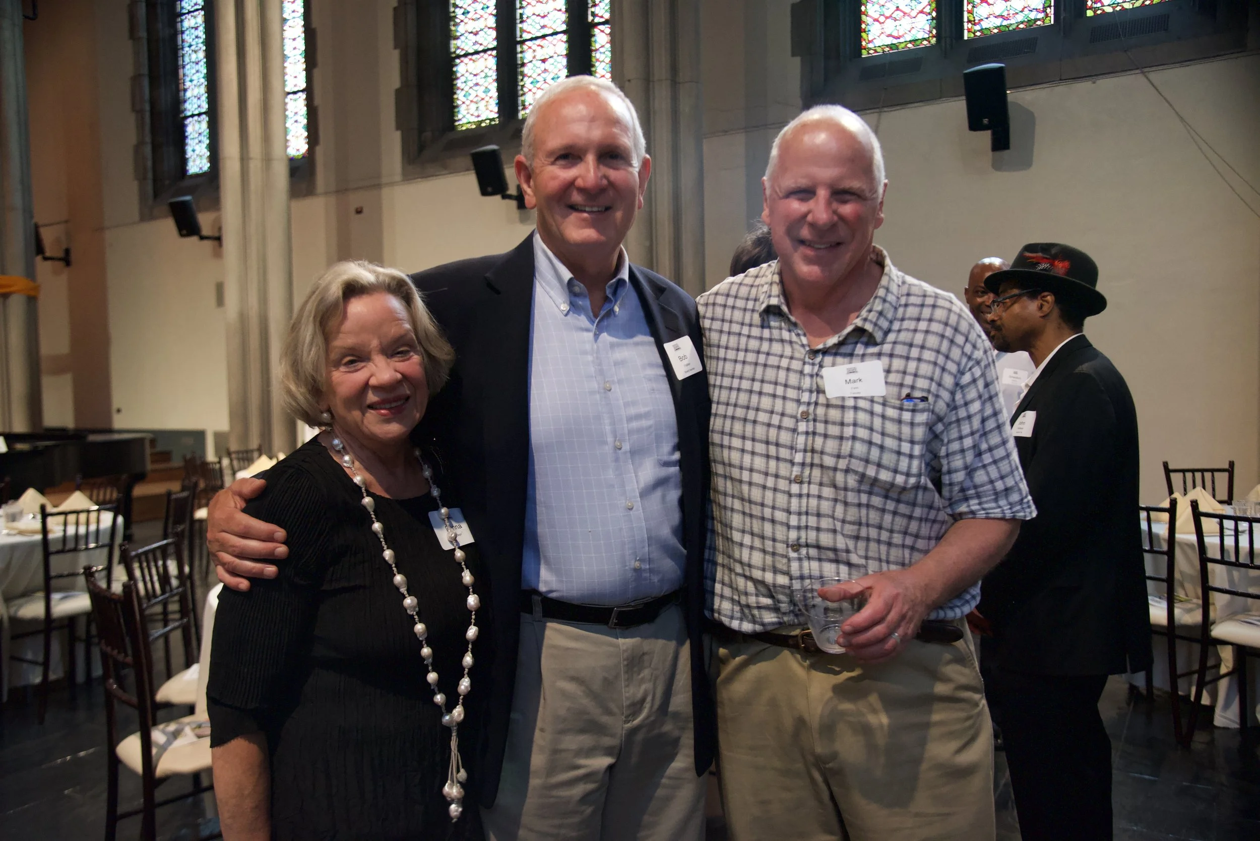 Three people smiling and posing for a photo at an event with stained glass windows in the background. They are dressed in semi-formal attire, with name tags on their shirts.