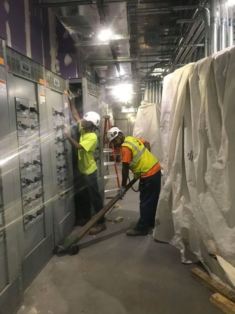 Two construction workers wearing safety helmets and high-visibility vests working with electrical panels in a utility or industrial room, with one using a tool and the other working on the panels.