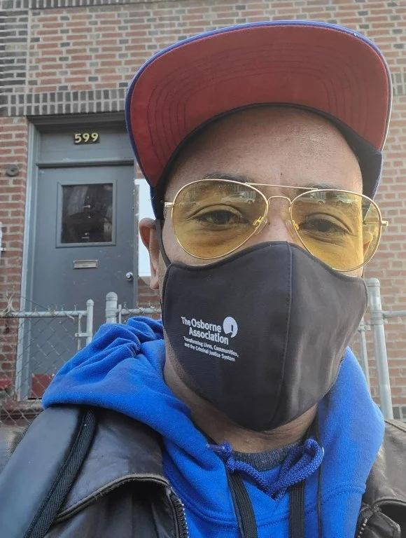Close-up of a man wearing a red and blue baseball cap, yellow-tinted glasses, a black face mask with 'The Osborne Association' logo, a blue hoodie, and a leather jacket, standing in front of a brick building with a gray door marked '599'.