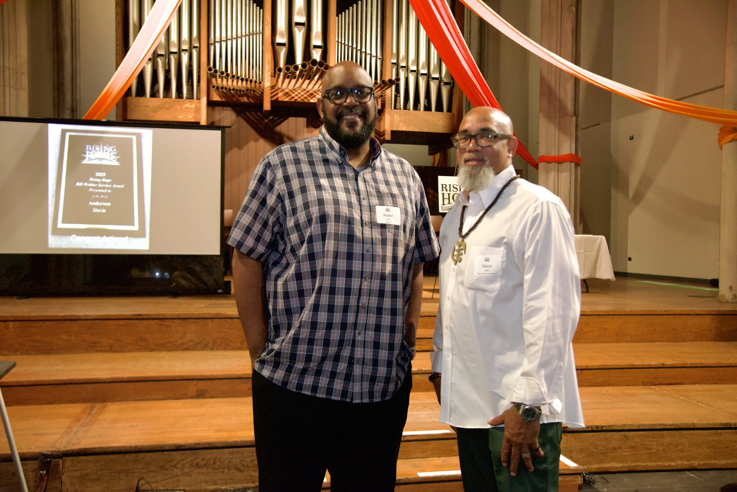Two men standing in front of a pipe organ with wooden scenery. One man is wearing a plaid shirt and glasses, and the other man is wearing a white shirt, glasses, and a beaded necklace. There is a presentation screen on the left with the words 'Rising