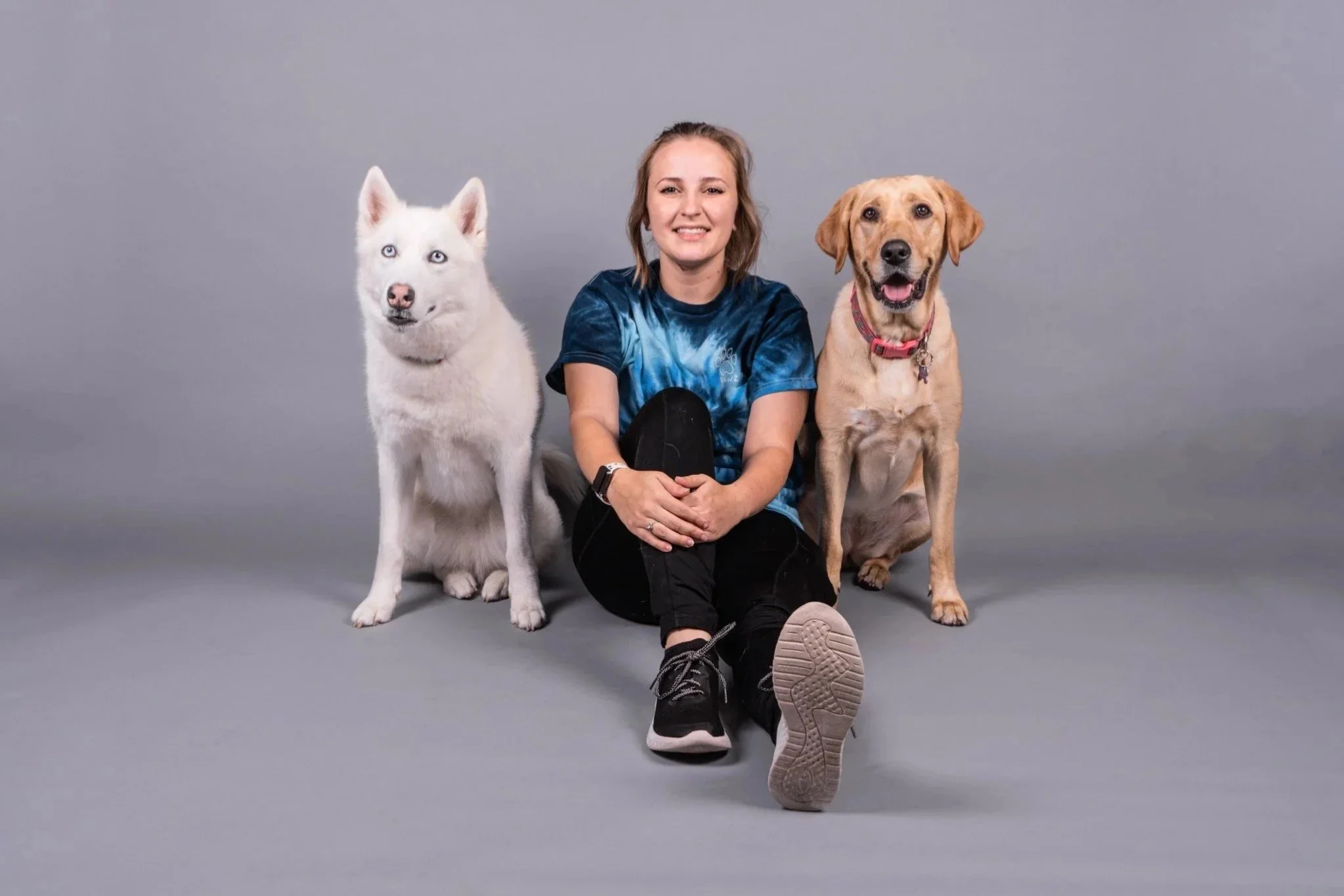 A young woman sitting on the floor with two dogs, one Siberian Husky and one yellow Labrador Retriever, against a plain gray background.
