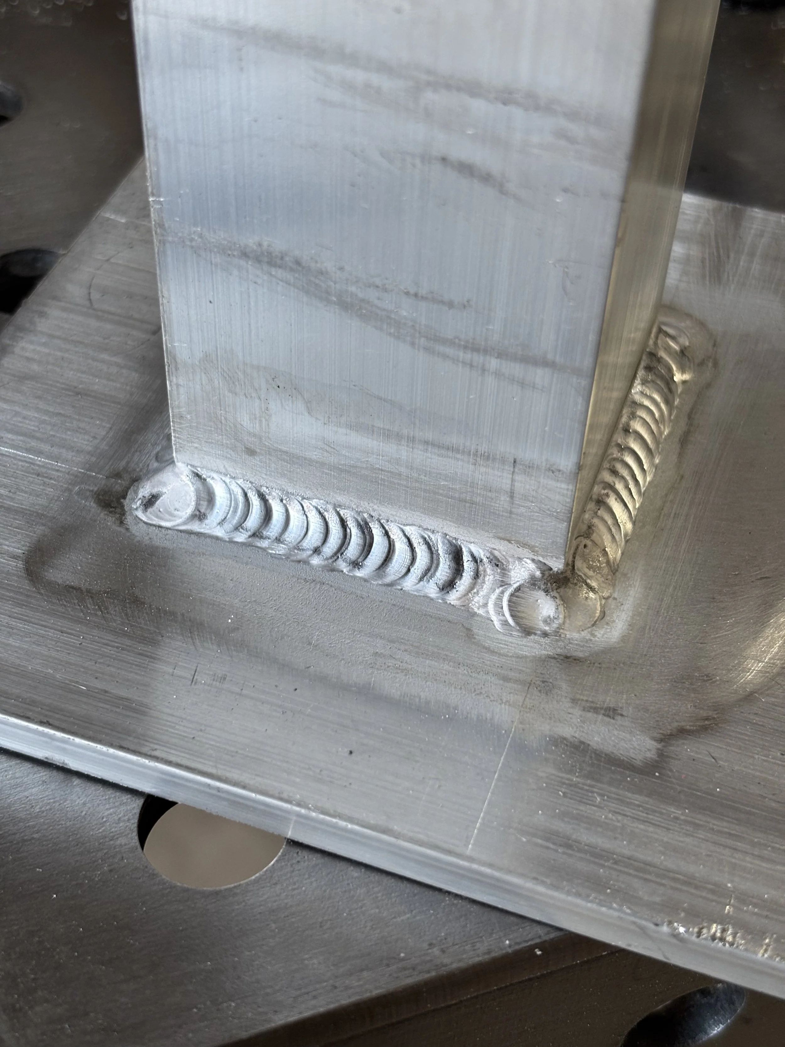 Close-up of a welded aluminum structure showing a weld bead along the edge, with a hole in the bottom left corner of the metal sheet.