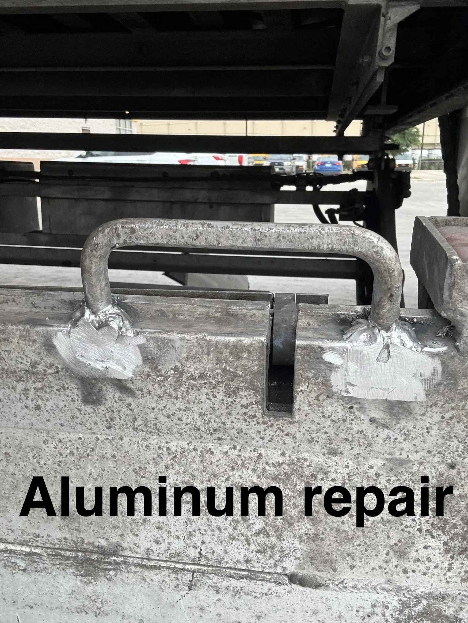 Underneath view of a metal structure with pipes and parts, with cars parked in the background, and the text 'Aluminum repair' written on the image.
