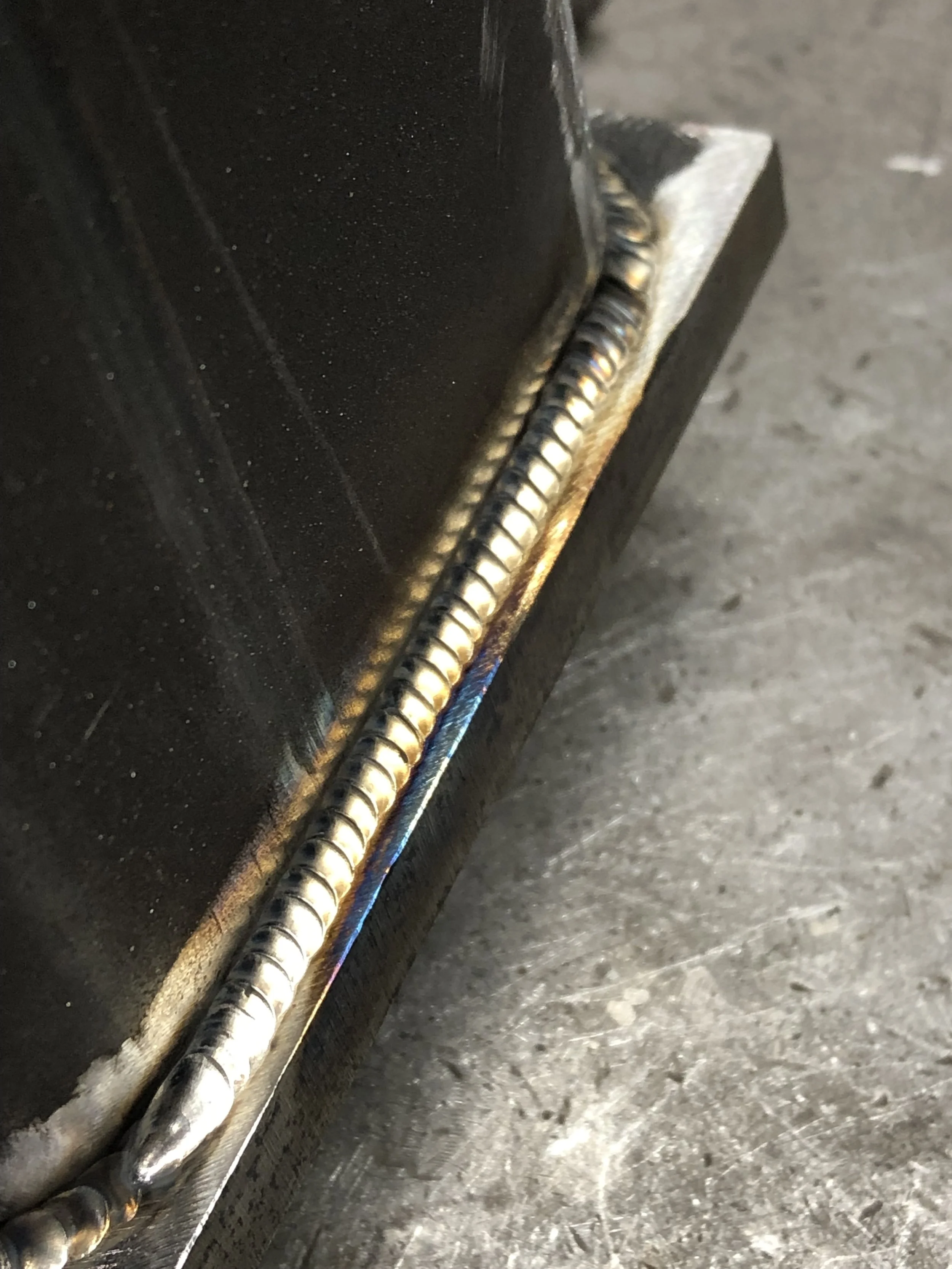 Close-up view of a welded metal corner joint, showing the weld bead along the edge of two black metal pieces with a rough gray surface beneath.