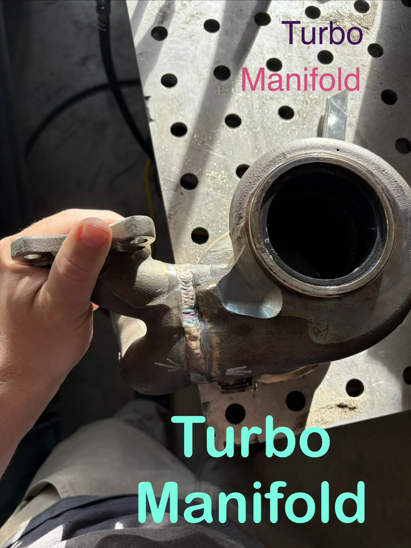 Close-up of a turbo manifold attached to an engine block with a person's hand in the foreground holding a small metal component.
