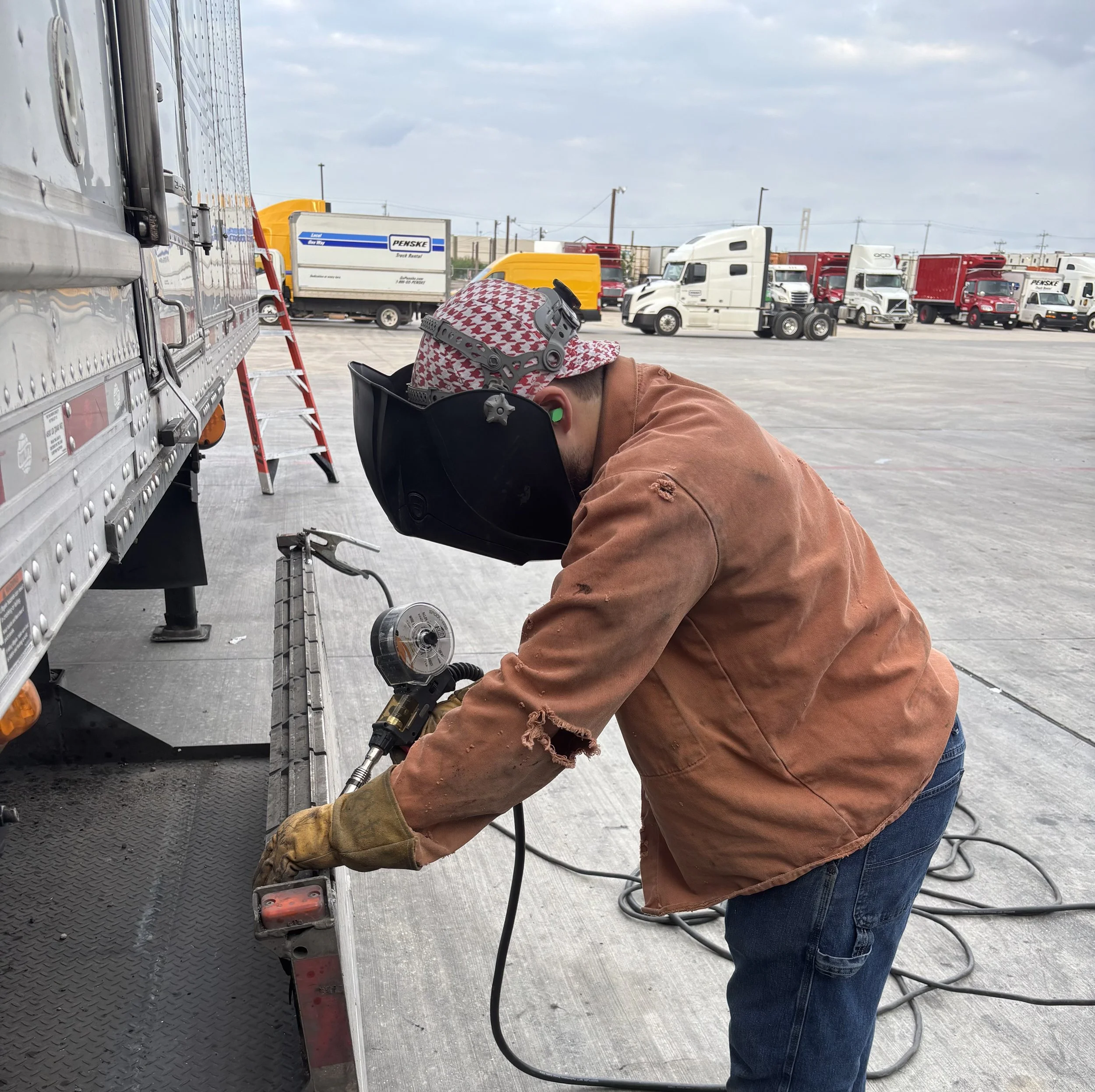 Worker welding or repairing a trailer in a truck yard, wearing a protective helmet and gloves.