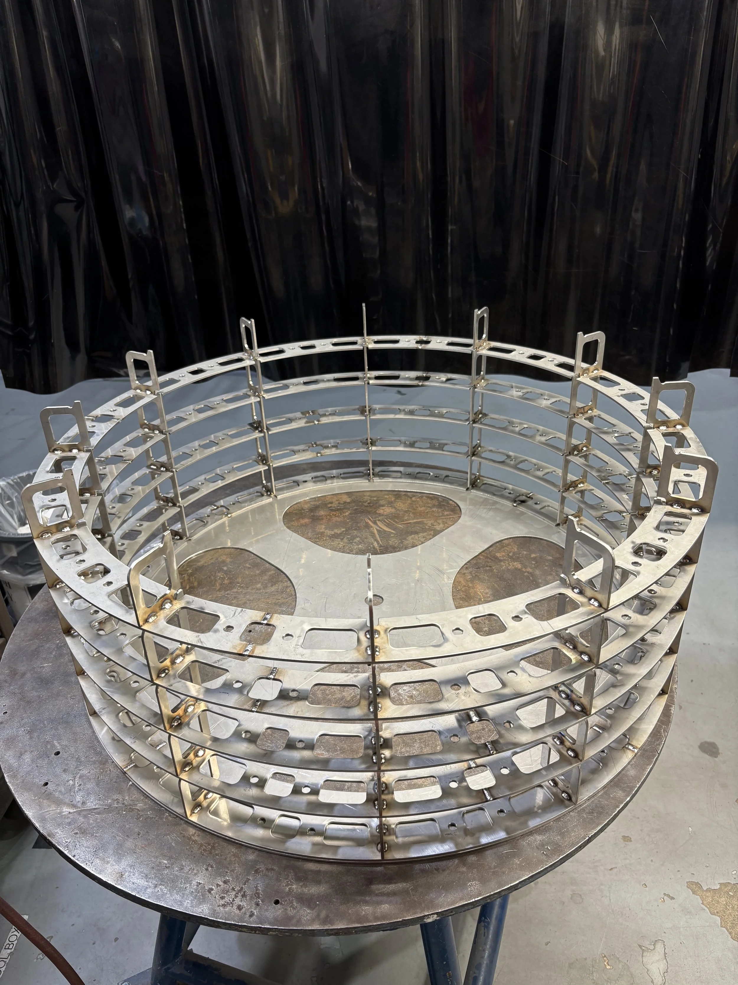 A circular metallic structure with multiple layered rings and numerous rectangular cutouts, placed on a worktable in a workshop.