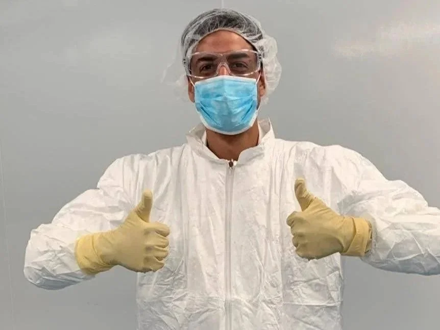 A person dressed in protective medical gear, including a white coverall, gloves, face mask, safety glasses, and a hair cap, giving two thumbs up.