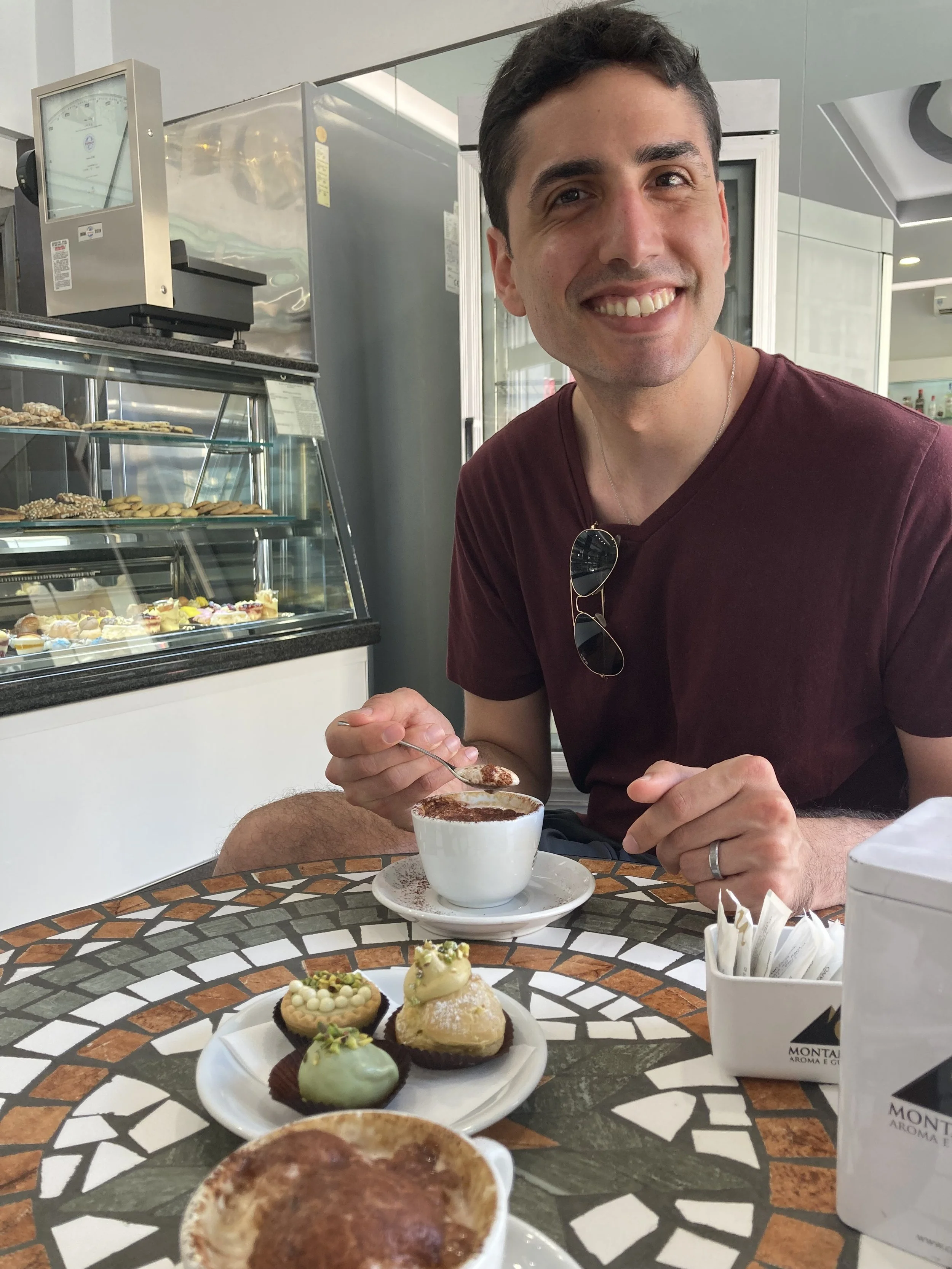 A man smiling while sitting at a table in a cafe, holding a spoon. On the table, there is a white plate with various decorated desserts, including cupcakes and pastries, and a cup of hot chocolate or coffee. In the background, there is a display case with more desserts and a young commercial oven.
