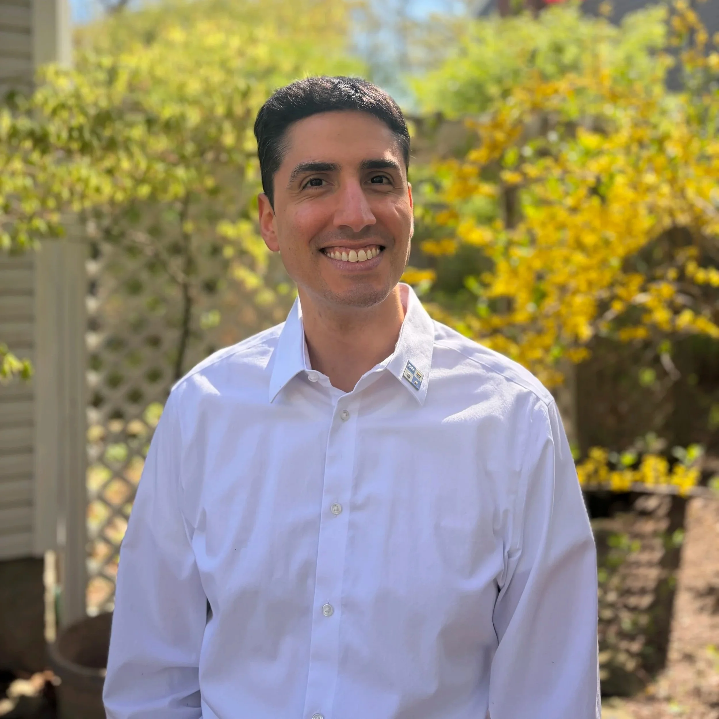 A smiling man wearing a white collared shirt standing outdoors with trees and fall foliage in the background.
