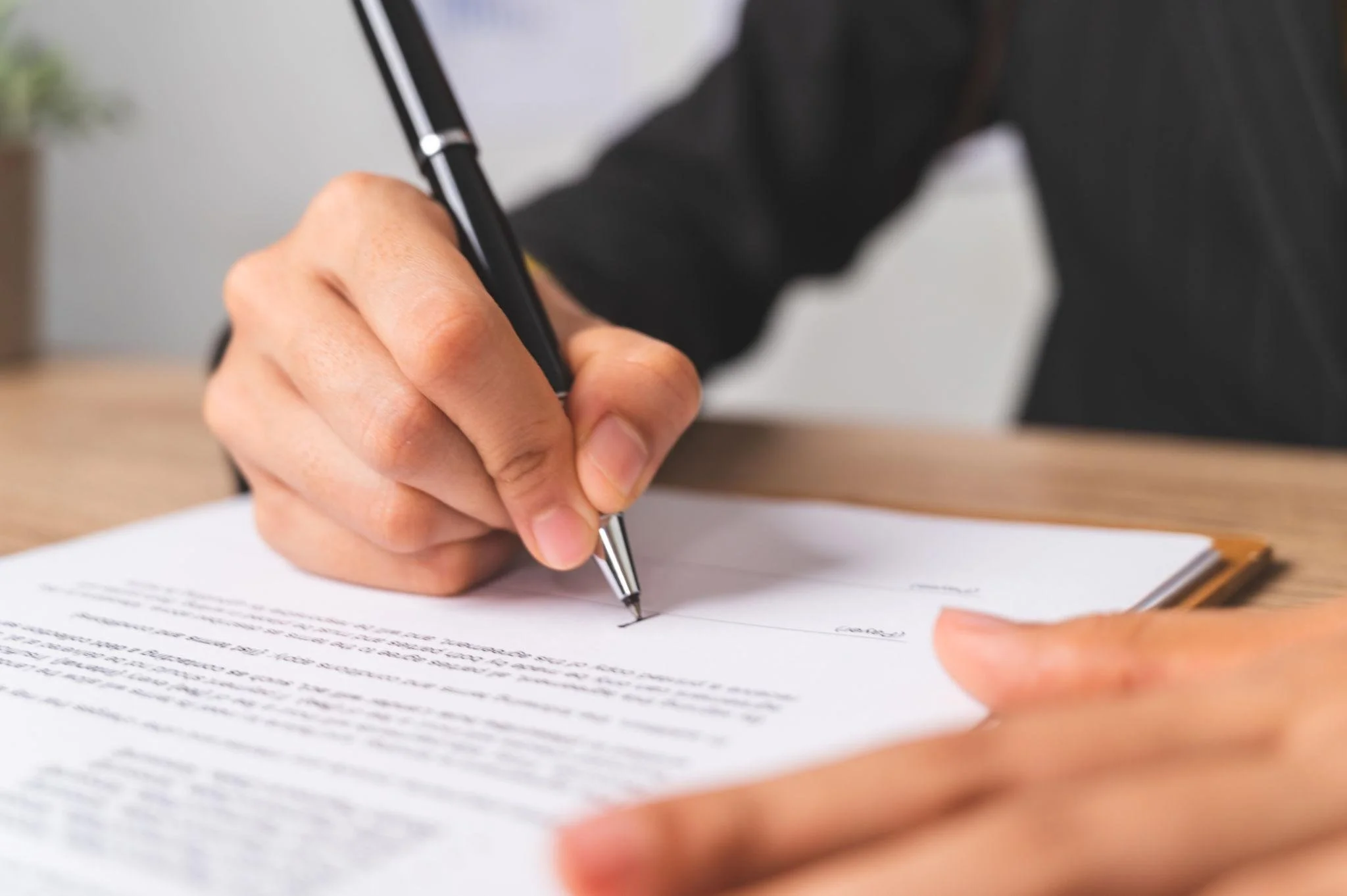 Close-up of a person signing a document with a black pen on a wooden desk.