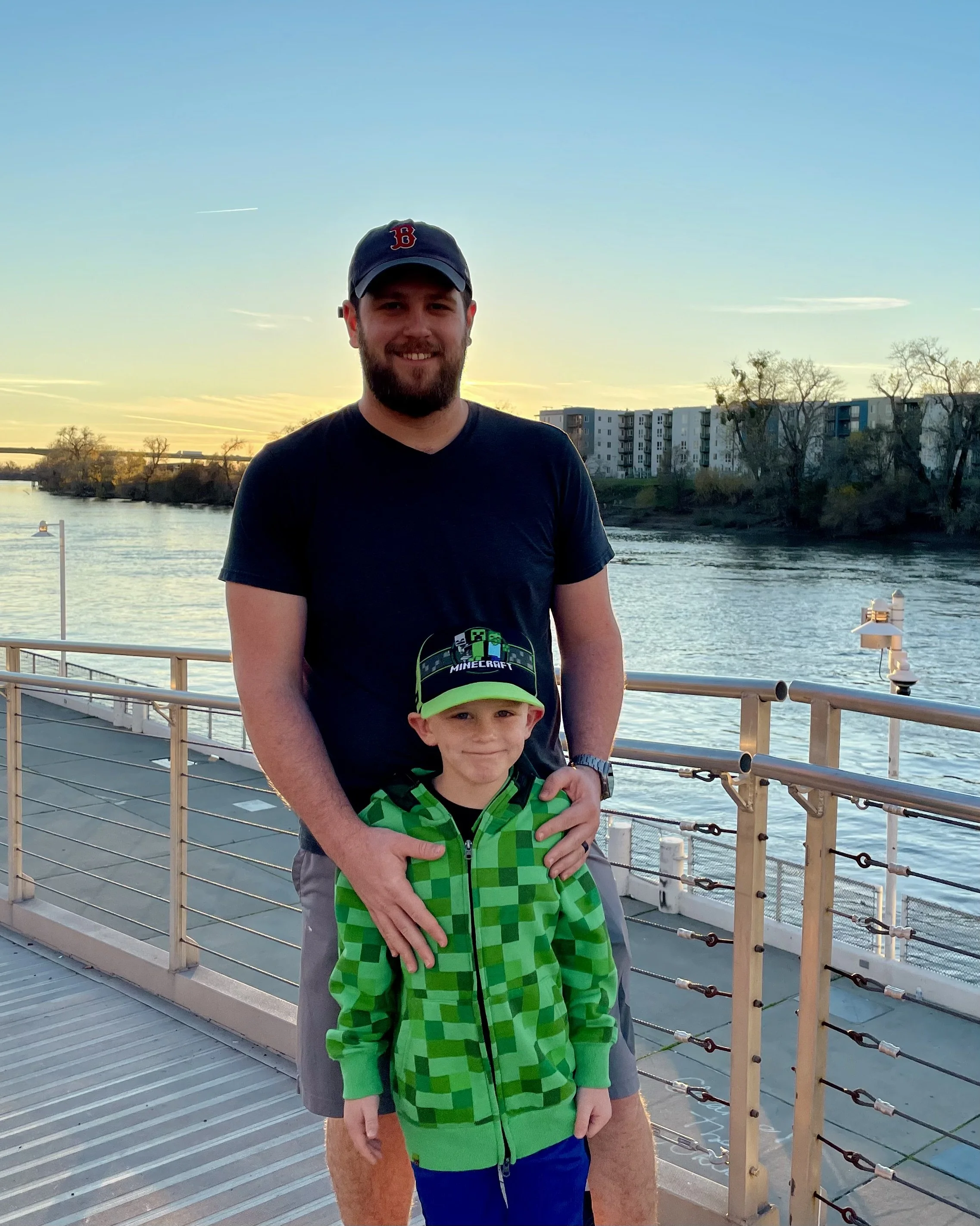 A man and a young boy standing on a boat dock at sunset with water and trees in the background.