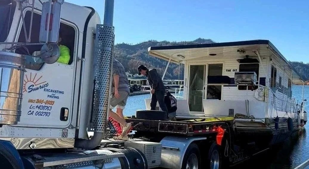 People loading or unloading a houseboat docked on a body of water, with mountains in the background.