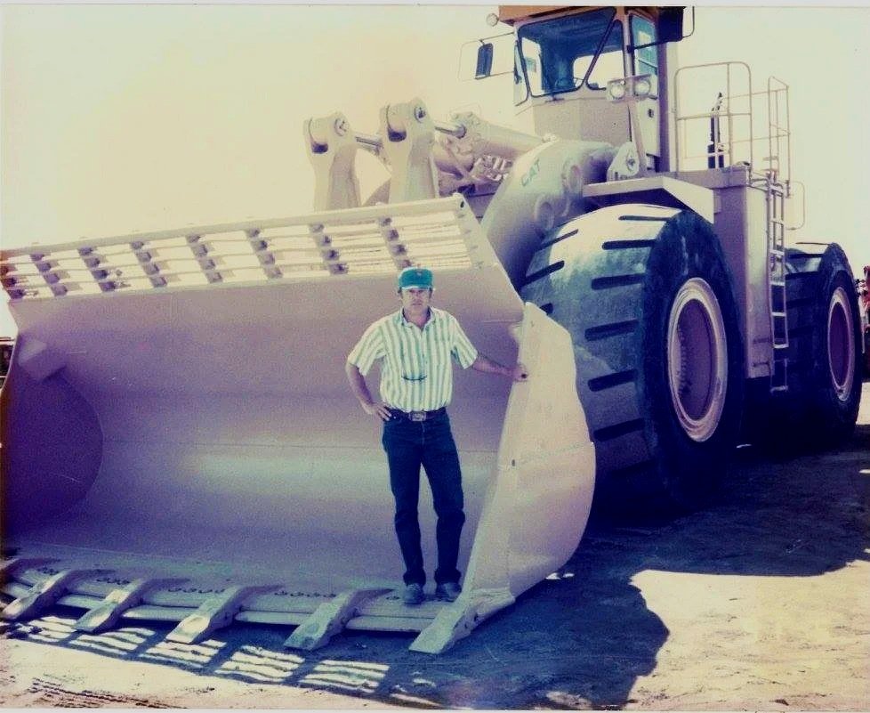 A man standing inside the large scoop of a construction tractor, with the tractor's massive tires in the background.