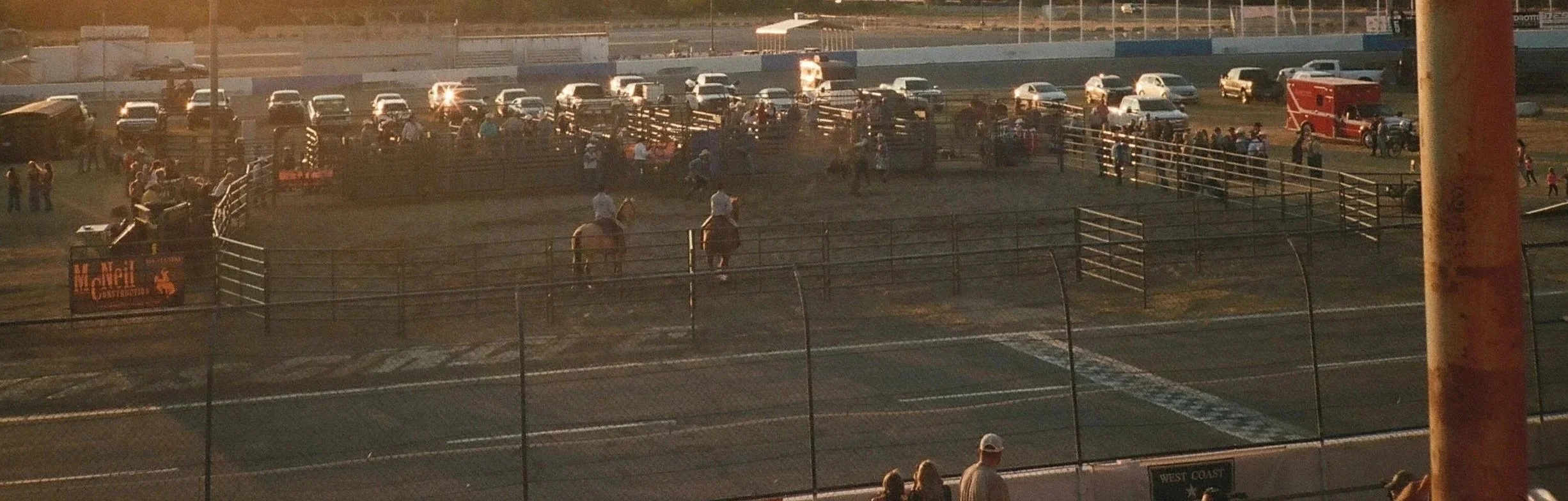Rodeo arena with cowboys on horseback and spectators watching. Cars are parked in the background, and the arena is enclosed by fencing.