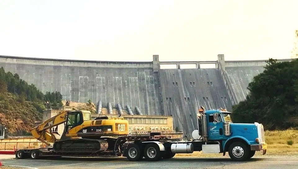 A large blue semi-truck carries a yellow Caterpillar excavator in front of a dam.