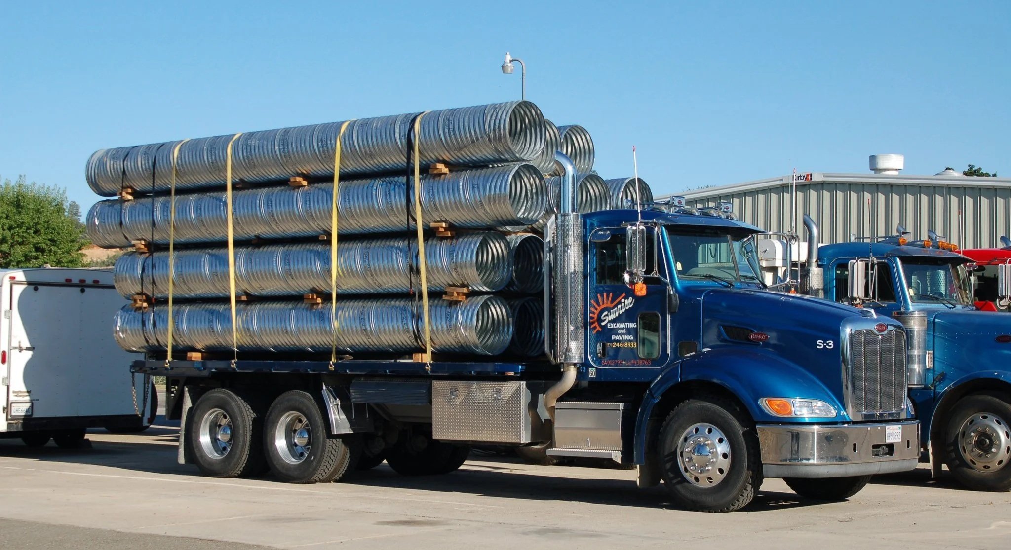 Blue semi-truck carrying multiple stacked metal ducts, parked in a lot near a metallic building with a clear blue sky.