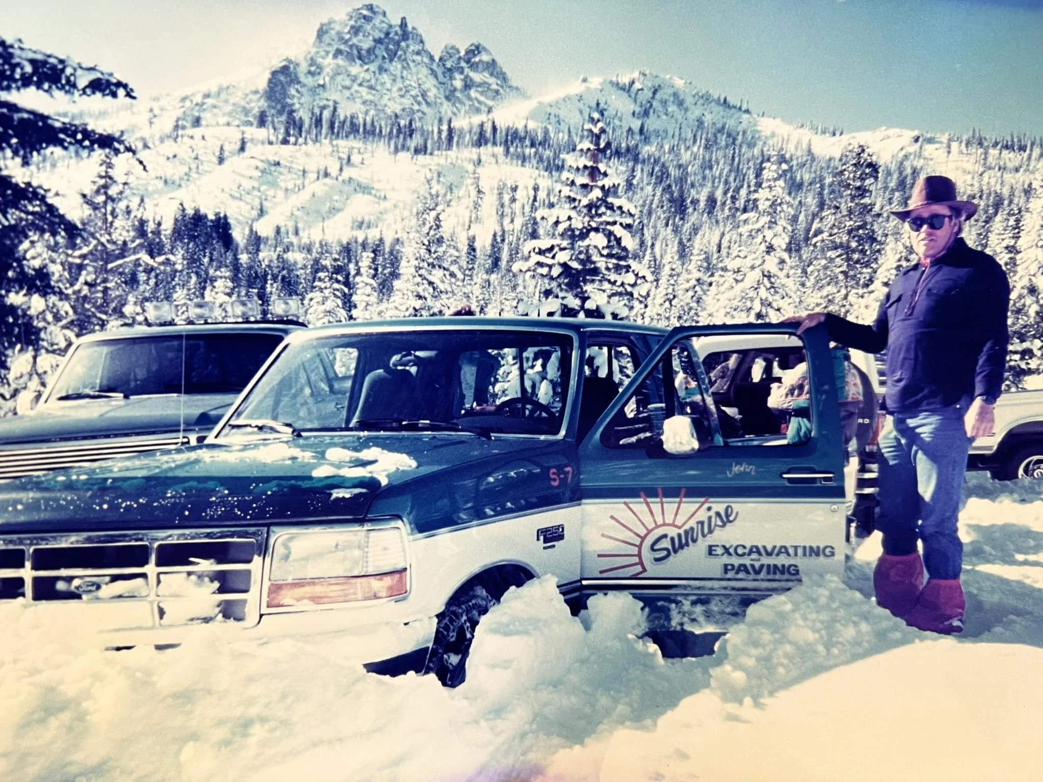 A man wearing sunglasses, a hat, and winter clothing stands next to a truck with a logo for Sunrise Excavating and Paving, parked in a snowy landscape with snow-covered trees and mountains in the background.
