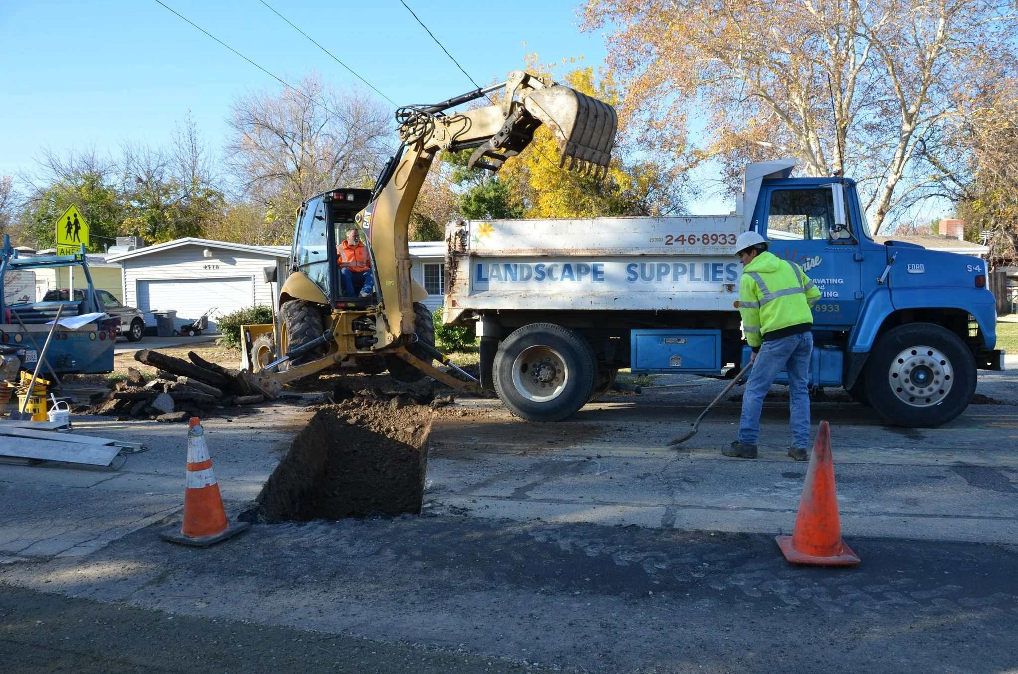 Construction workers operate machinery and work on a street excavation site, with orange cones placed around the area, in a neighborhood with houses and trees in the background.