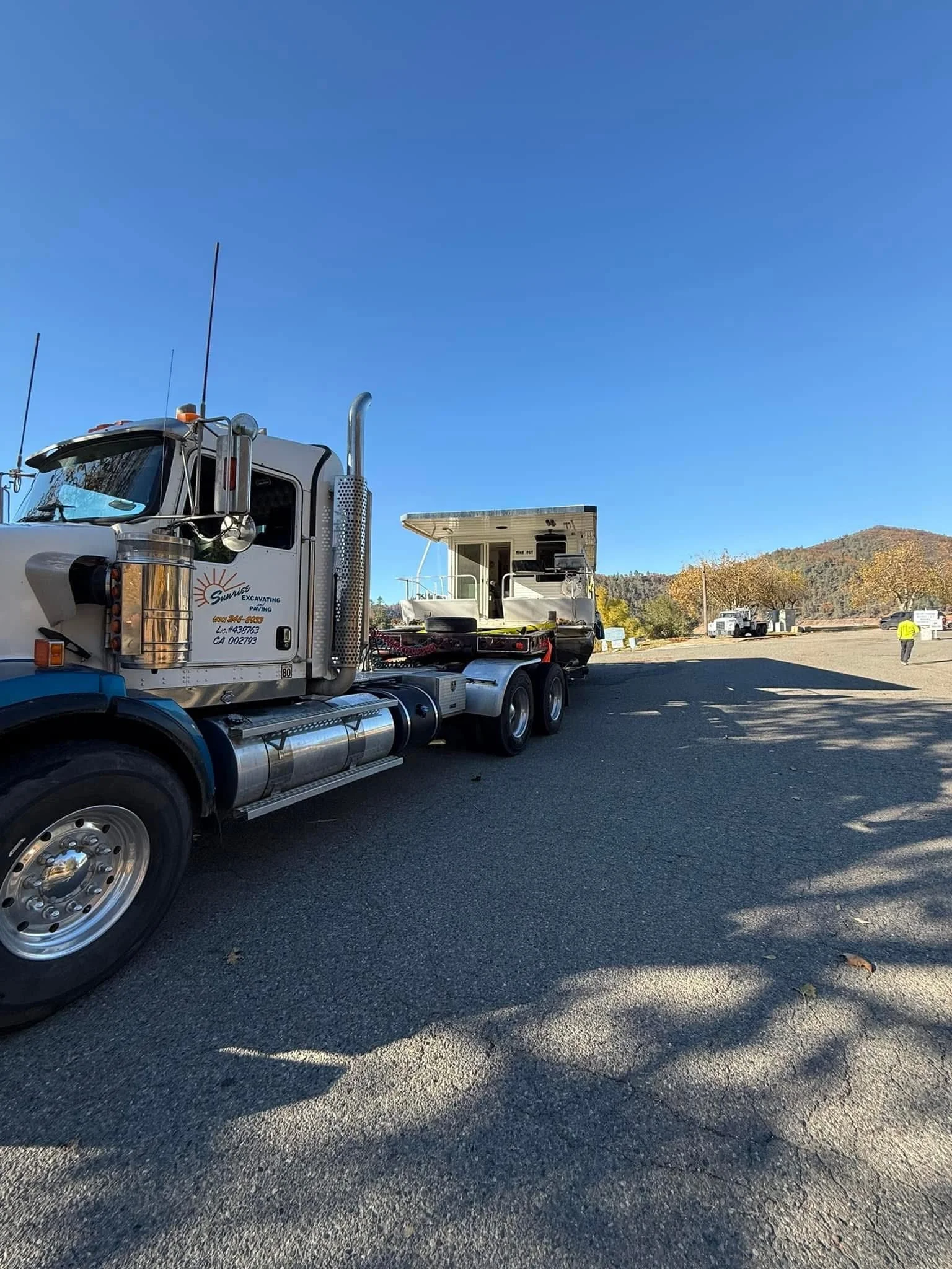 Semi-truck and trailer on a paved lot with a small white boat on the trailer and a person wearing a yellow safety vest in the background.