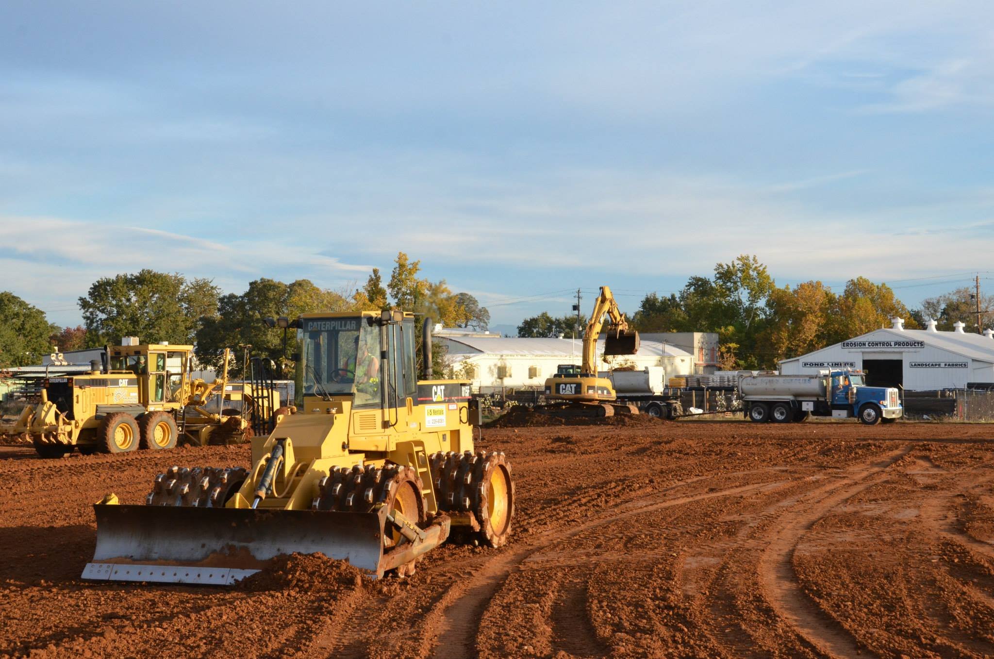 Construction site with yellow Caterpillar equipment, including a grader and an excavator, on reddish soil, with trees and industrial buildings in the background under a partly cloudy sky.