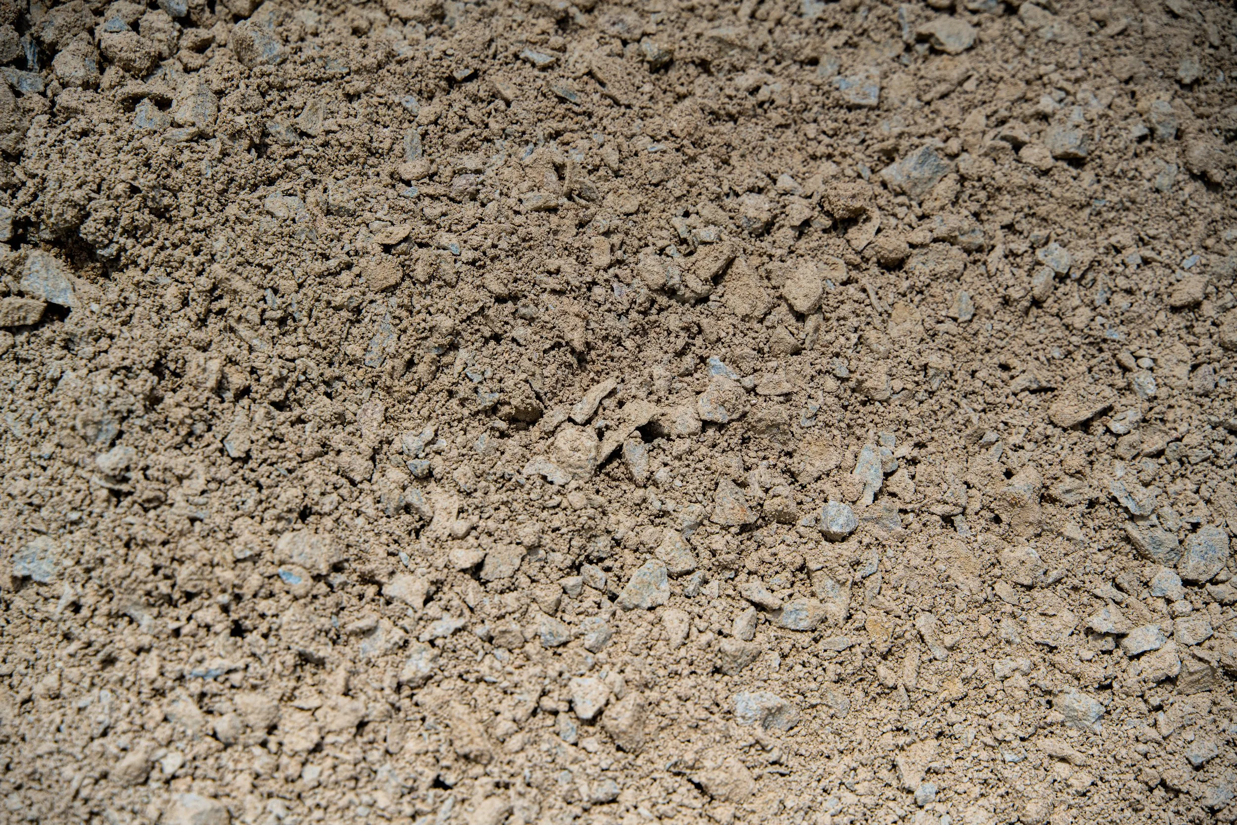 Close-up view of dry, loose, light brown dirt or soil with small rocks scattered throughout.