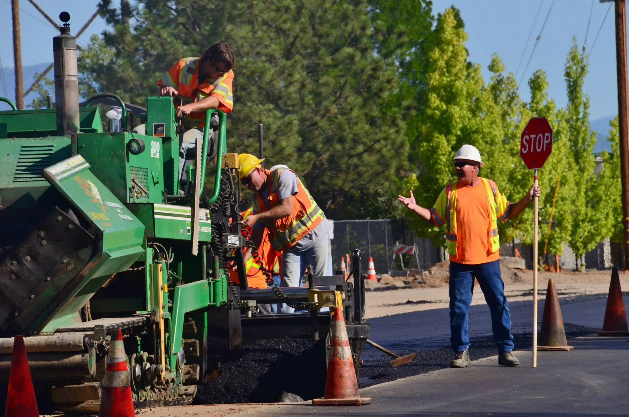 Construction workers wearing safety vests and helmets are operating a large green asphalt paving machine on a street. One worker is standing on the machine making adjustments, while another is examining equipment, and a third worker is holding a stop sign and gesturing to manage traffic. Orange cones line the area, and there are trees and utility poles in the background.