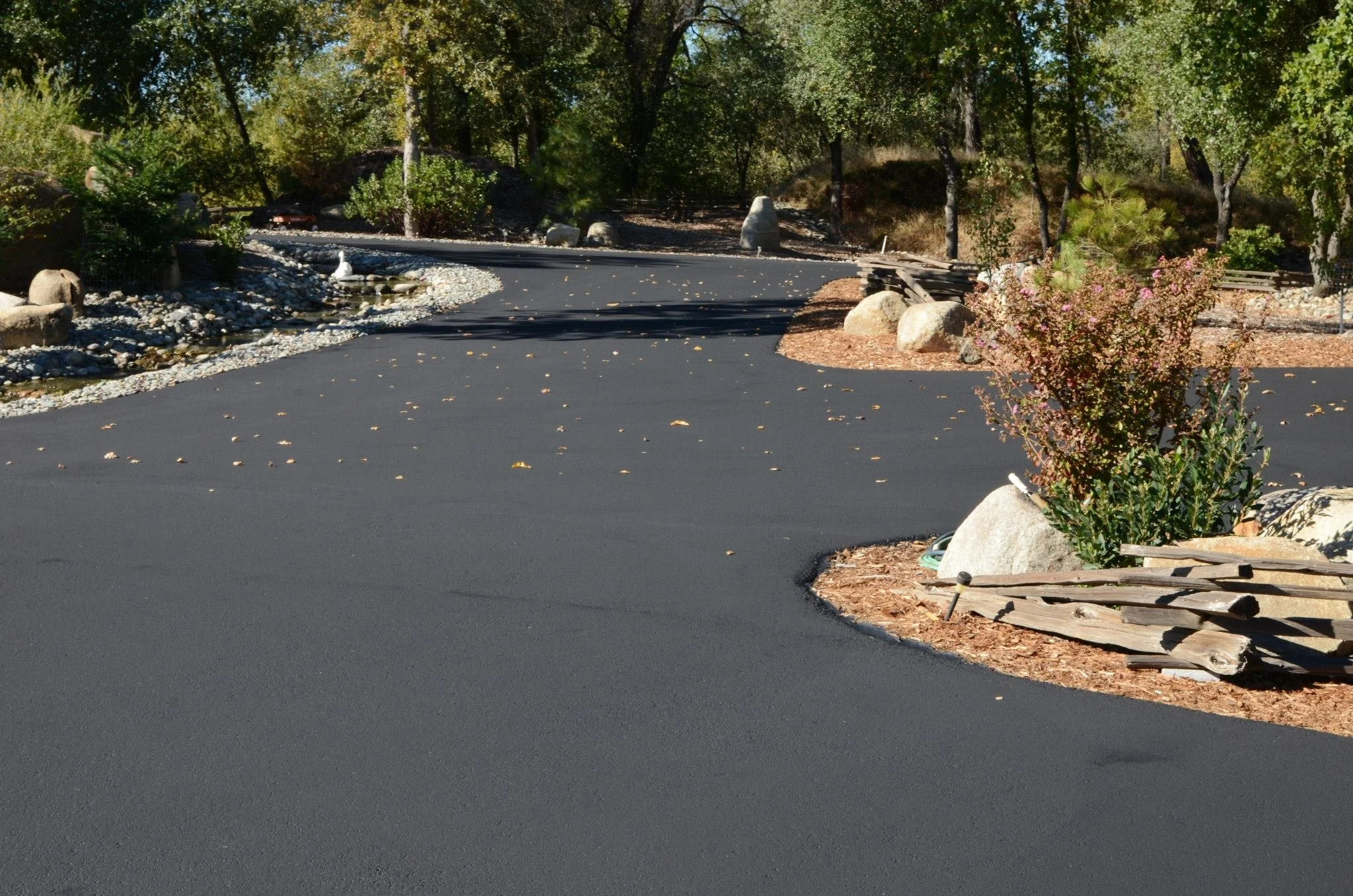 A paved driveway in a natural setting with trees, rocks, and a wooden fence on the right side. There is landscaping with plants and small rocks along the edges of the driveway.